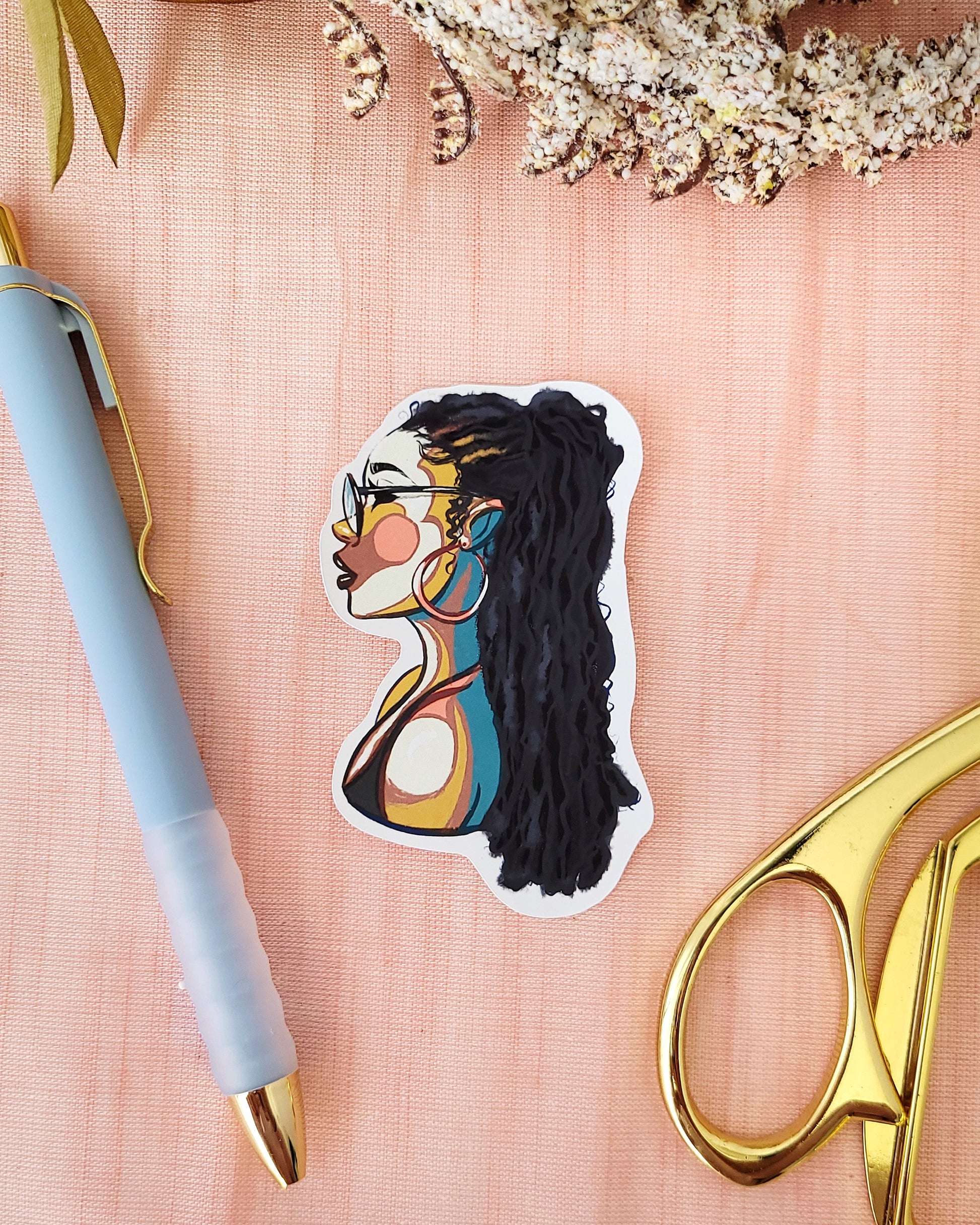 black woman with natural hair locs vinyl sticker illustrated my Gigi Moore of MoGigi. Resting on a soft pink fabric background with white flowers, blue ink pen, and cropped view of gold scissors.