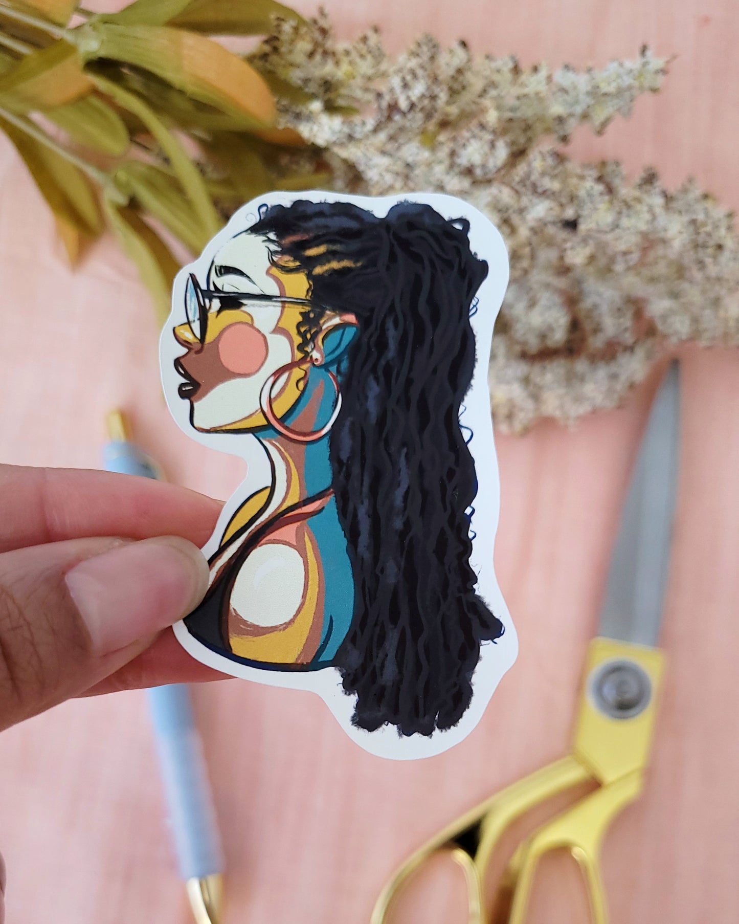 Close up view of black woman with natural hair locs illustration in a vinyl sticker with a soft pink background, with white flowers, blue gel ink pen, and pair of gold handled scissors. available on mogigi.shop from MoGigi bestsellers sticker collections.