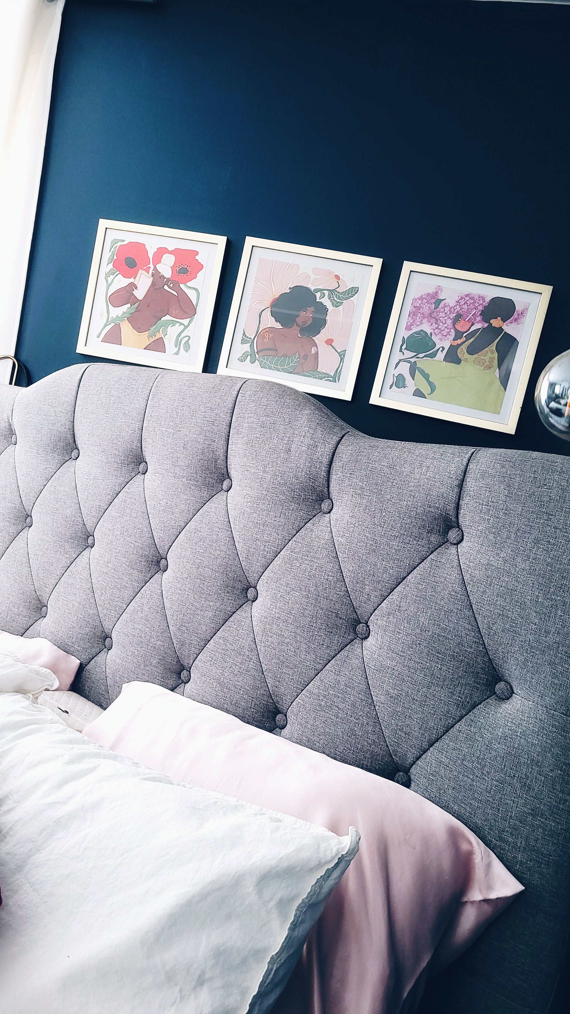 bedroom view showing black women gallery wall set hanging beyond the headboard. each 10 x 10 inch print are in a square gold finished picture frames. show up against a dark blue wall.