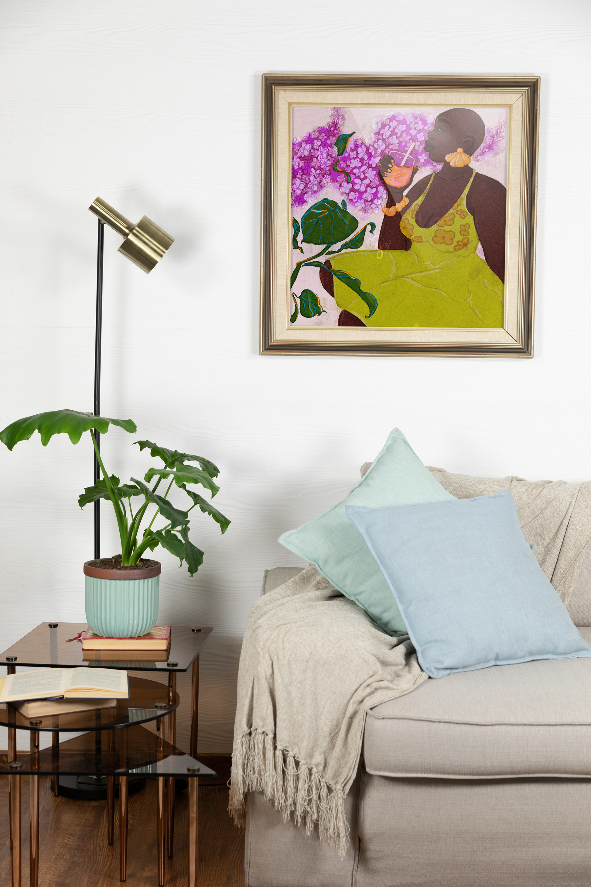 stylish black woman enjoy a summer picnic with lavender flowers around her. art print is in a gold frame hanging above the sofa. a small tiered side table is next to sofa with a floor lamp and potted houseplant.